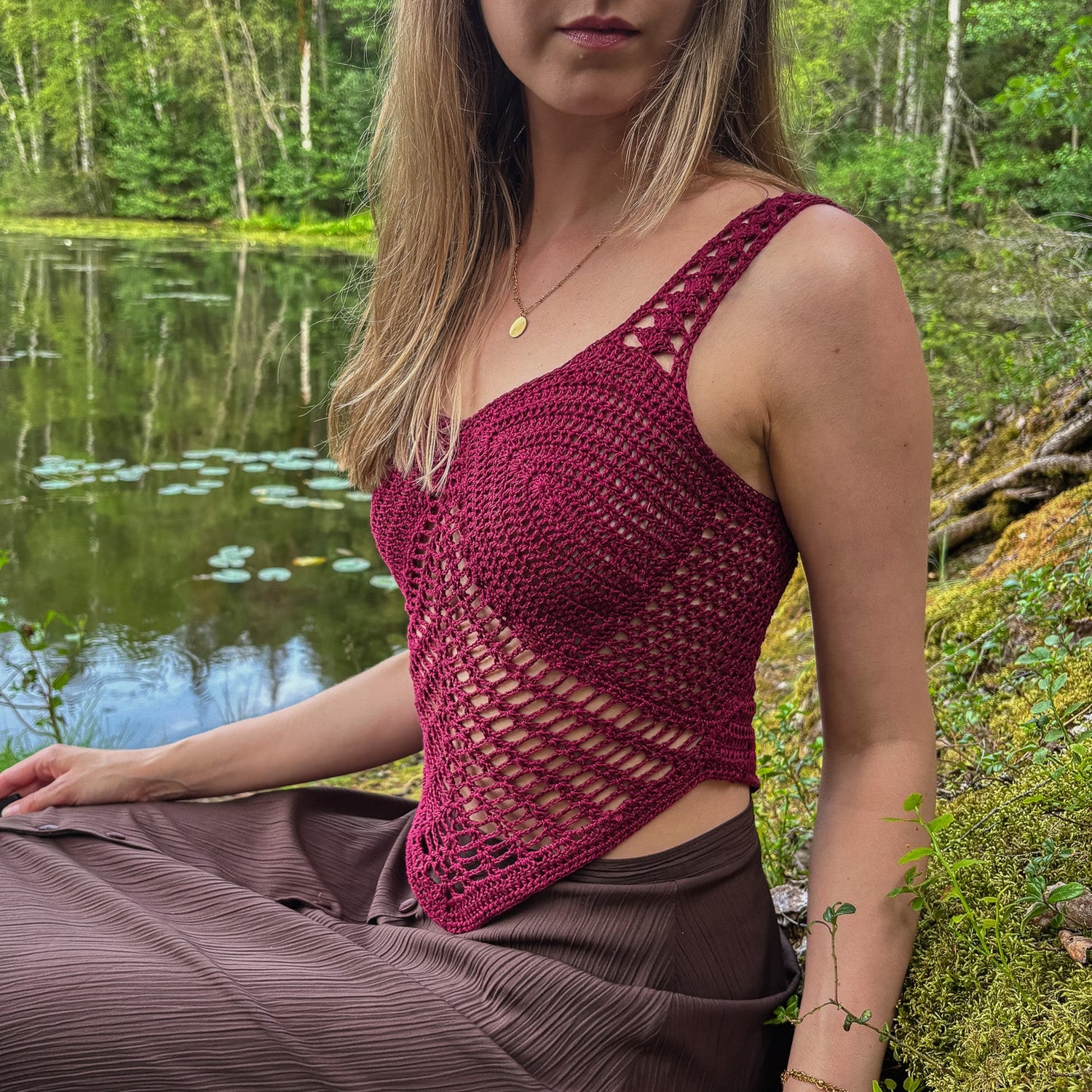 Woman wearing a maroon crochet top and brown pants sitting by a pond in a forest.